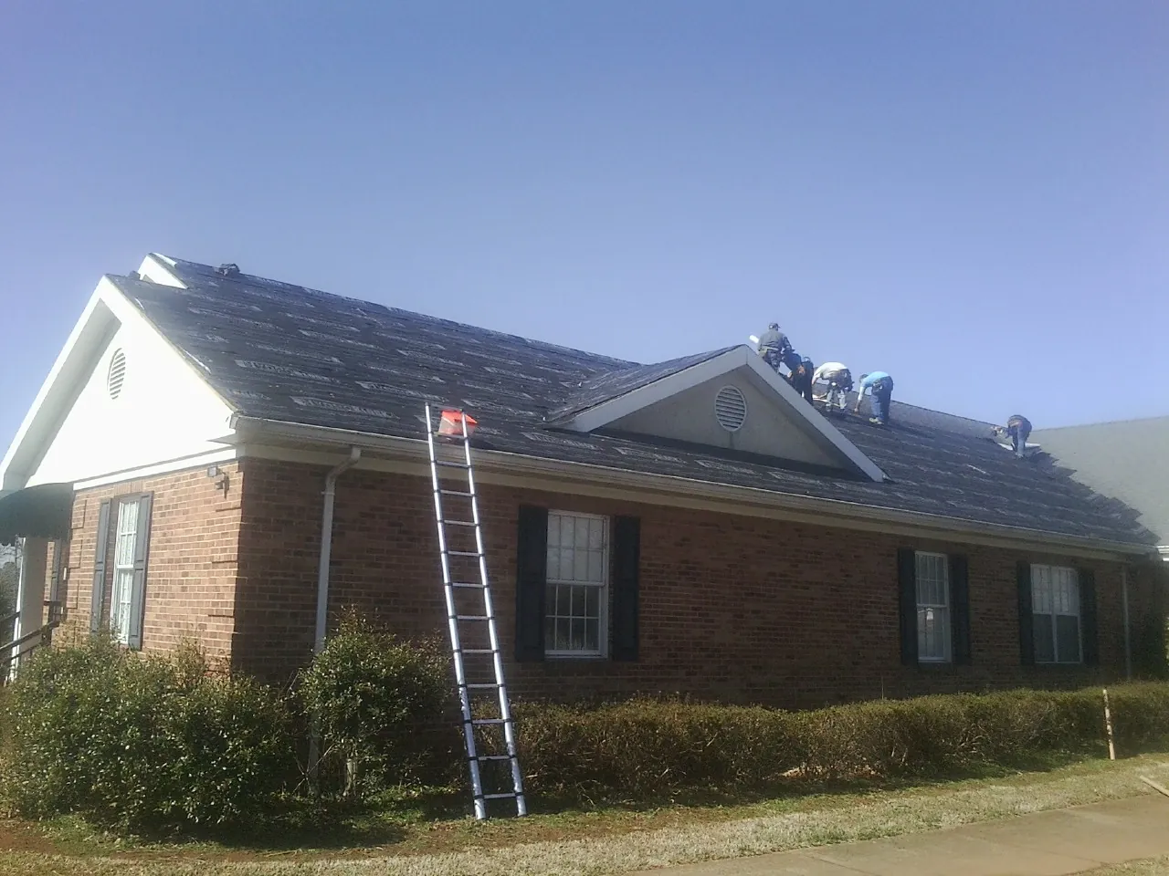 Crew tearing shingles off a brick ranch roof with a ladder set — Metal Roof Repair work by our crew in Baldwin